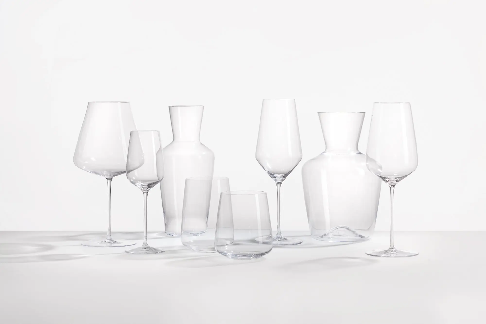 Vier wijnglazen op voet, twee karaffen en twee tumblers uit de Auriga-collectie naast elkaar op een witte ondergrond