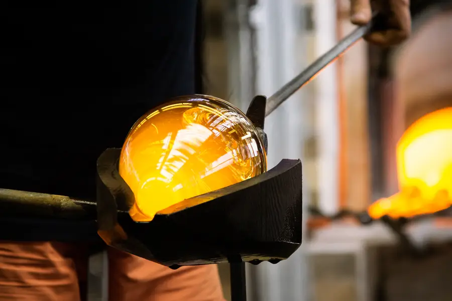 Een hand vormt met een tang de voet aan de steel van een glas, met de gloeiende oven wazig op de achtergrond